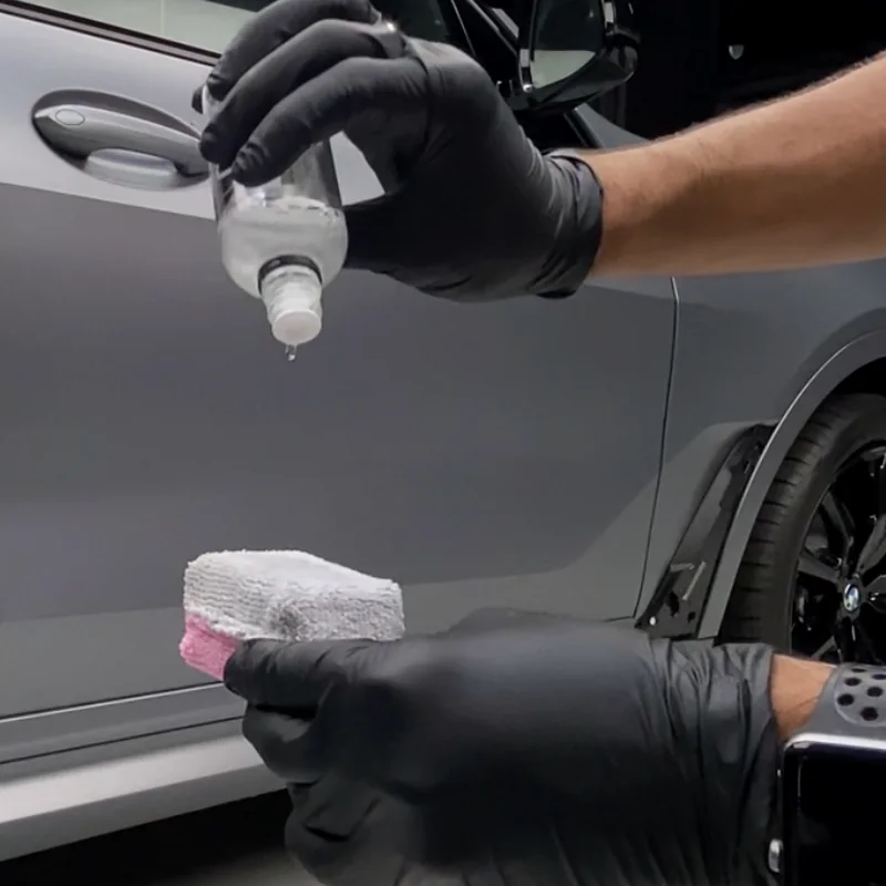 Gloved hands dosing ceramic coating from a dropper bottle onto a microfibre applicator beside a grey car